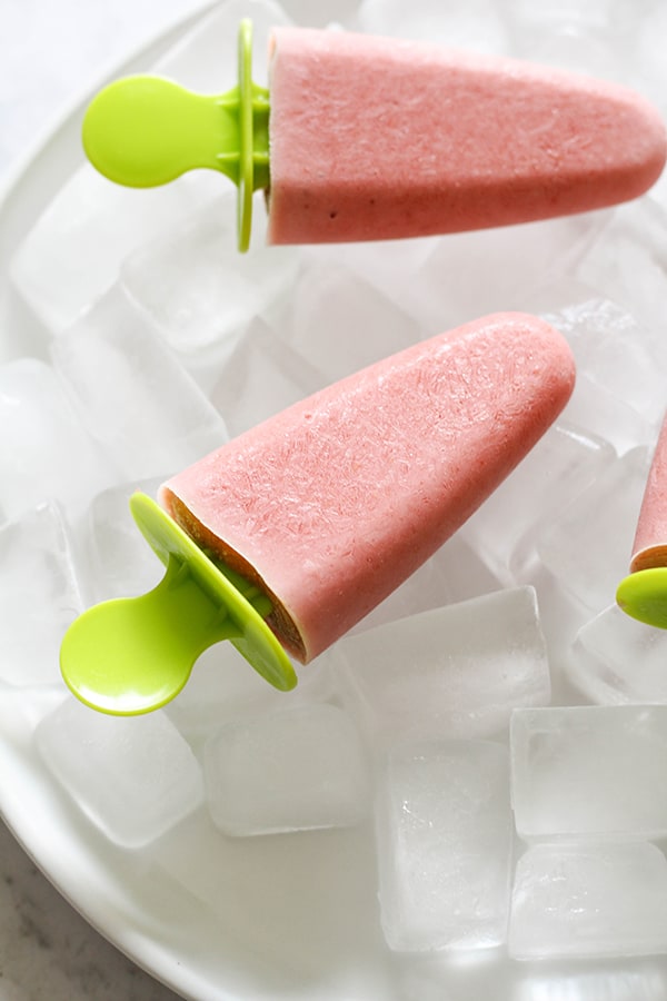 3 strawberry banana popsicles on a plate covered in ice cubes.