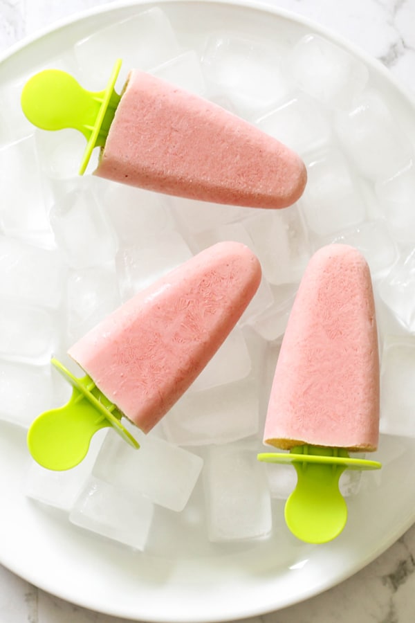 3 strawberry banana popsicles on a plate covered in ice cubes.