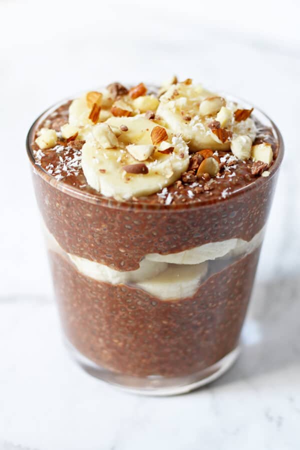chocolate chia pudding in small glass with banana slices, chopped almonds and coconut on top
