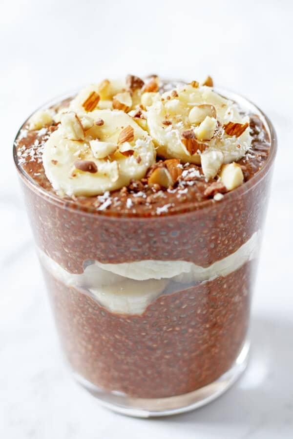 chocolate chia pudding in small glass with banana slices, chopped almonds and coconut on top