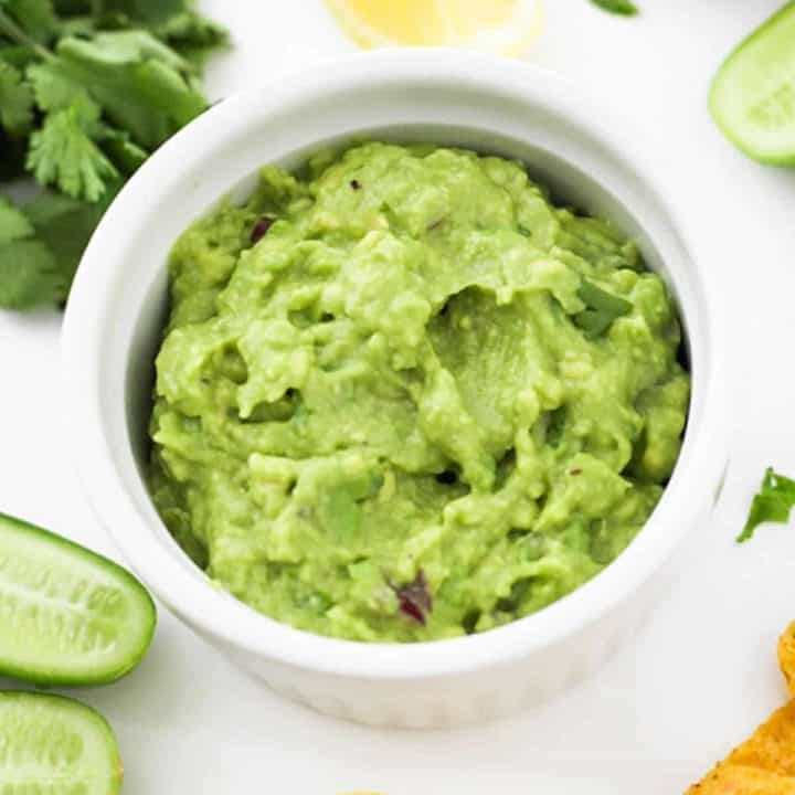 healthy guacamole in a white bowl surrounded by ingredients.