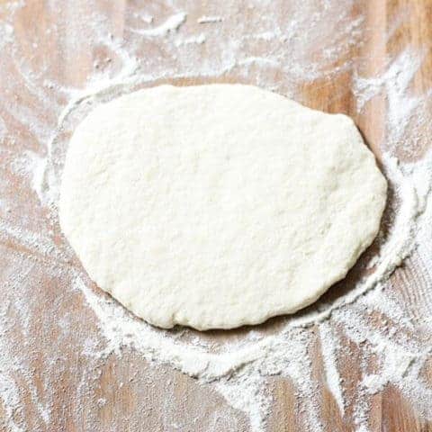 flattened pizza dough on a wooden cutting board.