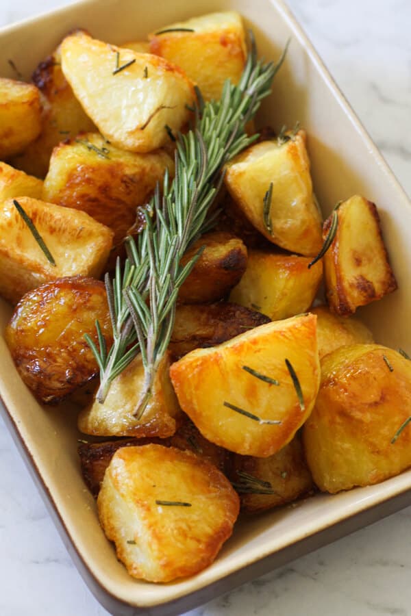 Crispy roast potatoes in a serving tray with rosemary sprigs on top.