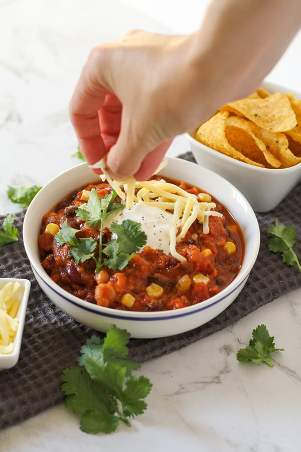a hand placing shredded cheese on a bowl of vegetarian three bean chili.