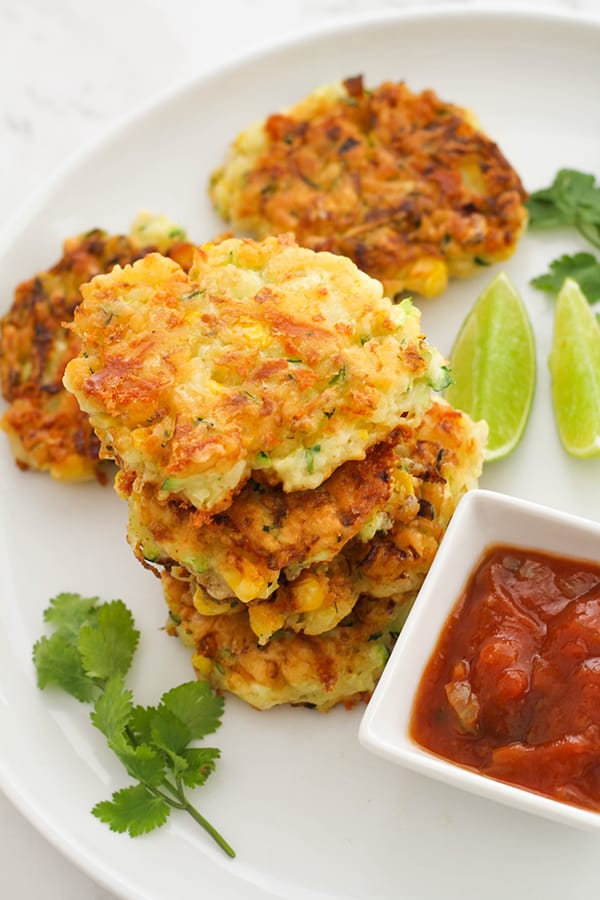 fritters stacked on top of each other on a white plate with salsa and limes.