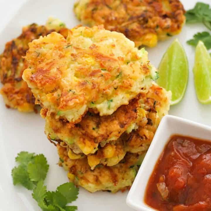 fritters stacked on top of each other on a white plate with salsa and limes.